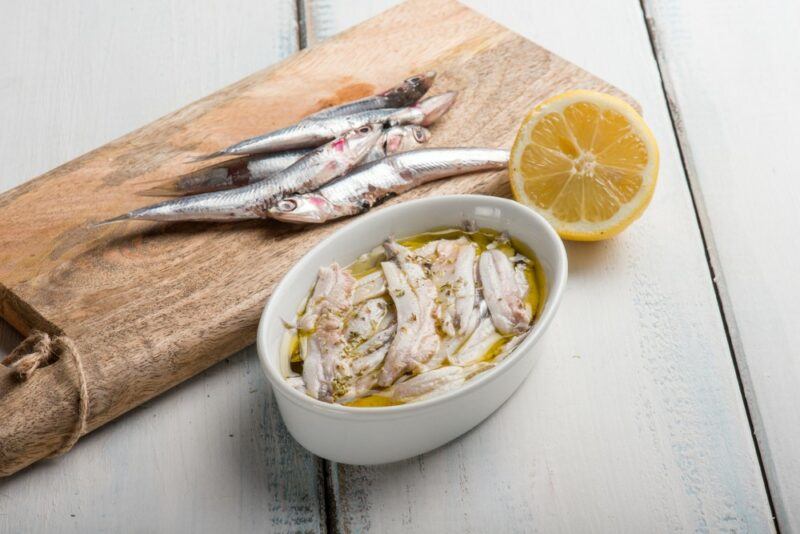 A wooden board with a couple of fresh anchovies, next to a white dish containing anchovies and oil