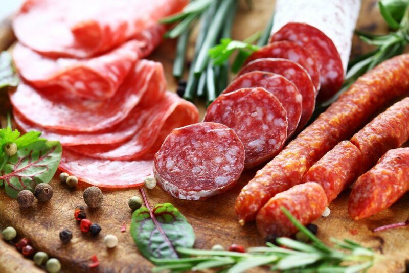 A wooden board with thick and thin slices of salami, plus three cured sausages. There are fresh herbs and peppercorns on the board as well.