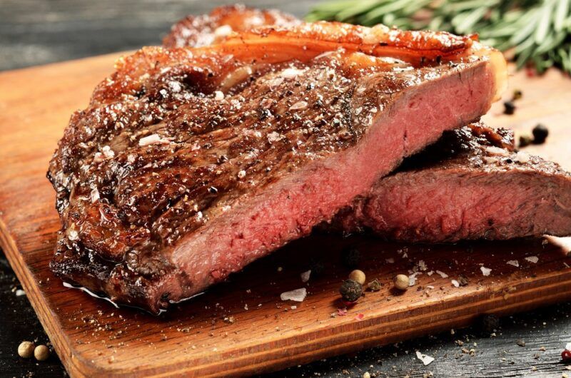 A wooden board with a large piece of steak with cracked pepper and rosemary