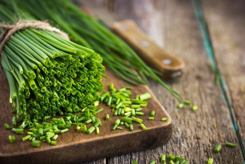 A wooden board with a large bundle of chives, where someone has started to chop them. There's a knife next to the board, on a wooden table