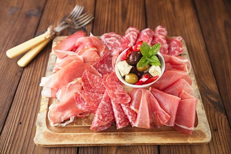 A wooden tray containing a selection of cold cut meat with a small bowl of olives and feta