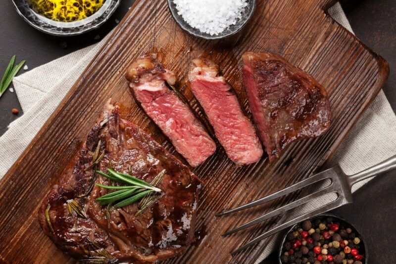 A wooden board with a large piece of cooked steak, where two small slices and one large chunk have been cut off. There's also a bowl of salt and one of peppercorns, plus a fork, in the image