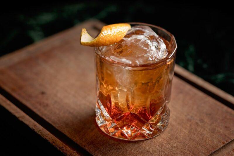 A single old fashioned cocktail with ice and a lemon twist, on a wooden board