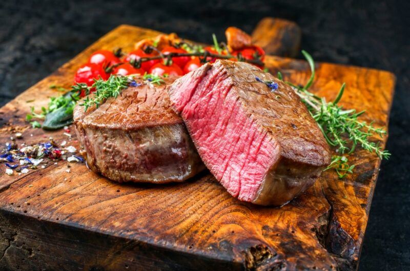 A large wooden board with a filet mignon steak that's been cut in half, next to fresh herbs and cherry tomatoes