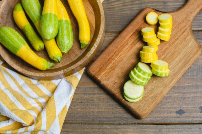 A wooden board with sliced Zephyr squash, next to a plate that contains whole zephyr squashes. The yellow and green gradient can be clearly seen on the squash