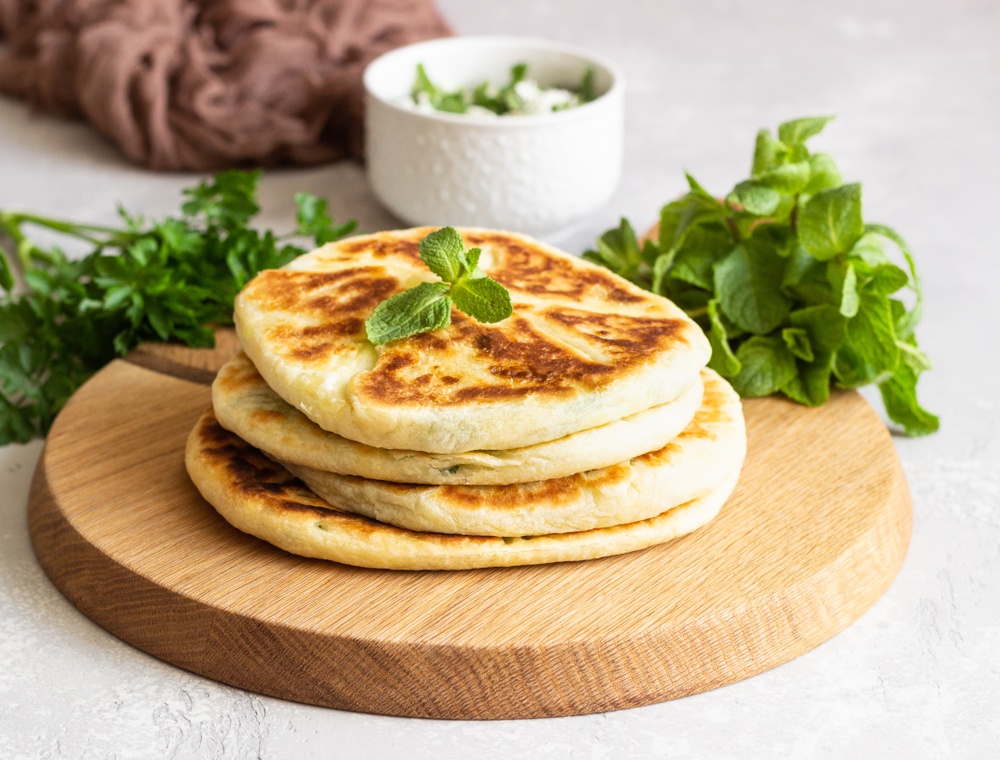 A wooden board with a stack of flatbread, some greens, and a white container of dip