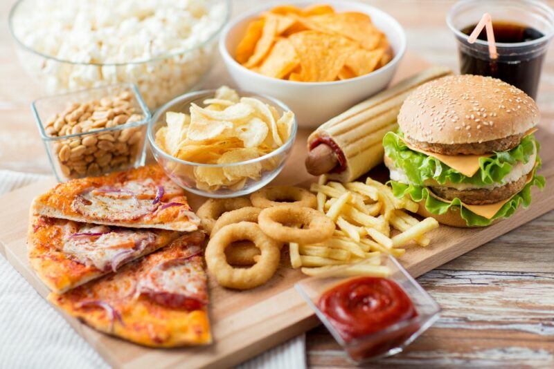 A wooden board with fast food, including a burger, pizza, onion rings, cola, and other fast food items.