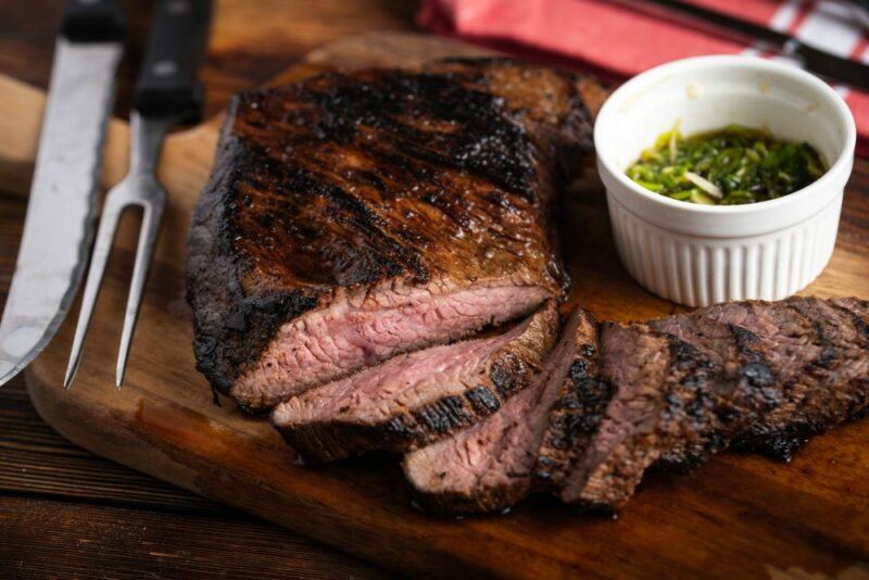 A wooden board with a tri tip steak that's been well cooked and sliced into thin pieces, next to a white dish of green sauce and carving tools