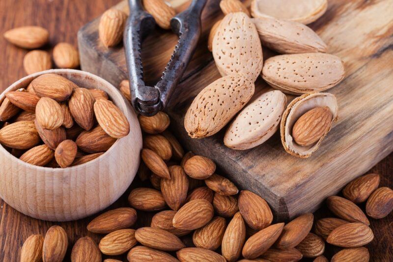 A bowl of almonds, with more almonds on the table, and a wooden board with almonds in their shell