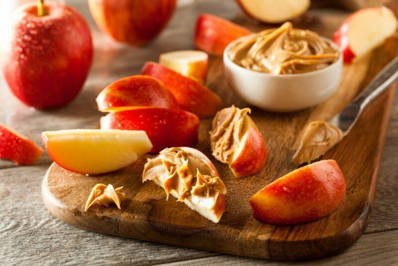 A wooden board with wedges of apple and a small bowl of peanut butter. Some of the apples have peanut butter spread onto them.