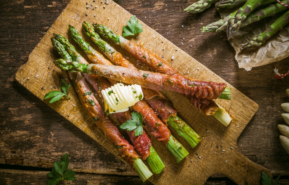 A wooden board with pieces of bacon wrapped asparagus and some butter