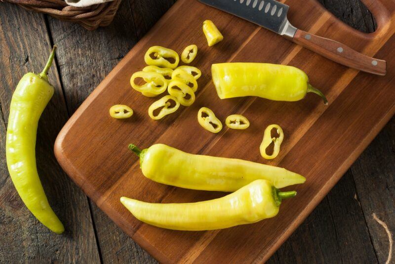 A wooden board with two whole banana peppers, a banana pepper half, and sliced banana peppers. There's a single banana pepper next to the board