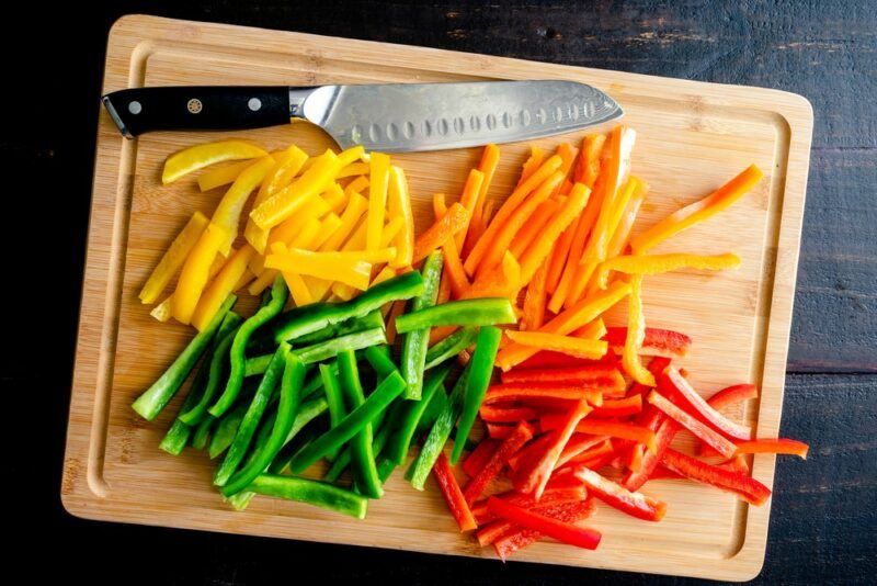 A wooden chopping board with sliced yellow peppers, orange peppers, green peppers, and red peppers, plus a knife