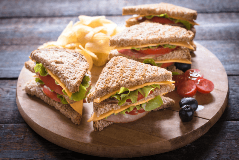 A club sandwich that has been cut into four sections, on a board with chips, olives, and tomatoes. 