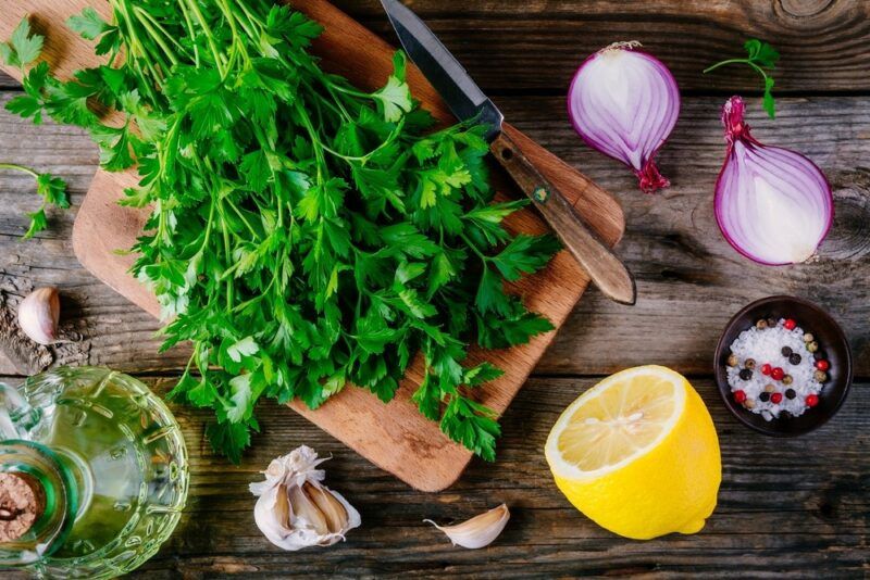 A wooden board with fresh parsley, with other fresh ingredients scattered around, including onions, salt, and garlic