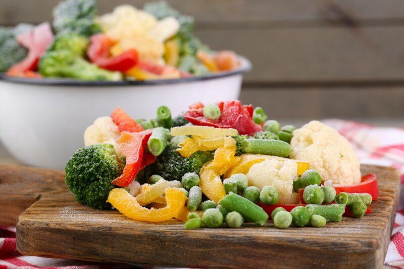 A wooden board with frozen vegetables, in front of a large white and blue bowl that contains even more of them