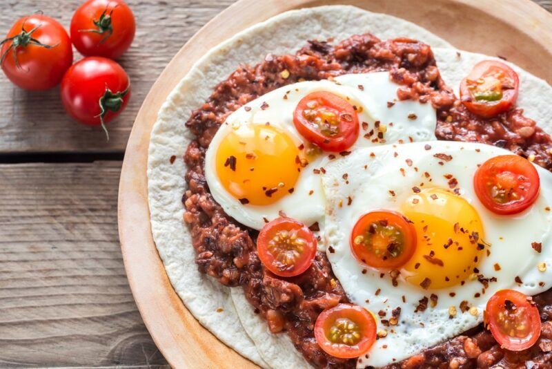 A wooden board with a dish called huevos rancheros that includes tortillas, beans, eggs, and tomatoes