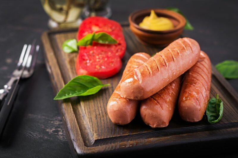 A wooden board with four plant-based sausages, a small container of mustard and some sliced tomatoes