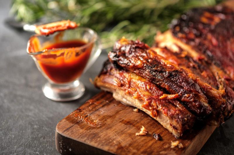 A wooden board containing pork ribs covered in glaze, next to a glass jug of glaze