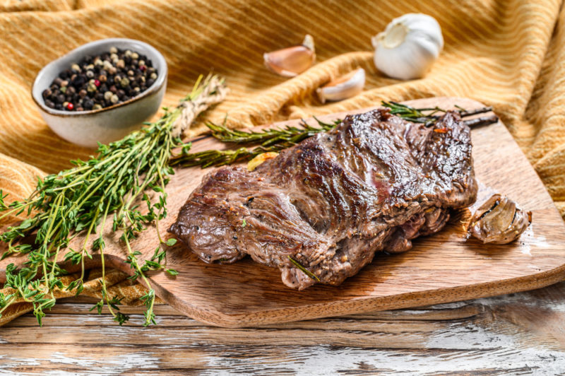 A wooden board with a piece of cooked red meat, next to a sprig of herbs and some seasonings