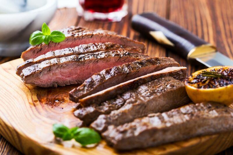 A wooden board with sliced flank steak next to a knife and some herbs