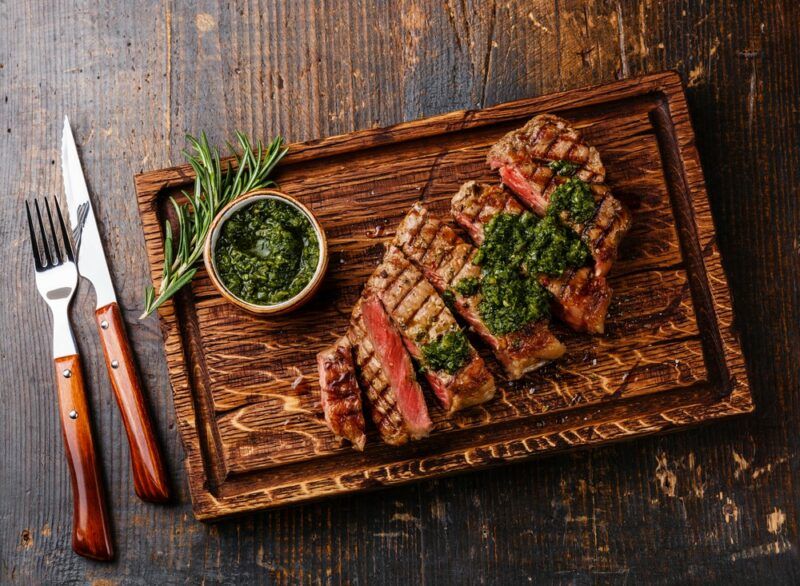 A wooden table with a wooden board containing a piece of sliced sirloin steak, some herbs, and green sauce, next to a knife and fork