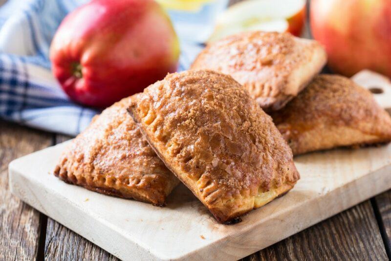 A wooden or marble board containing small apple pies with apples in the background