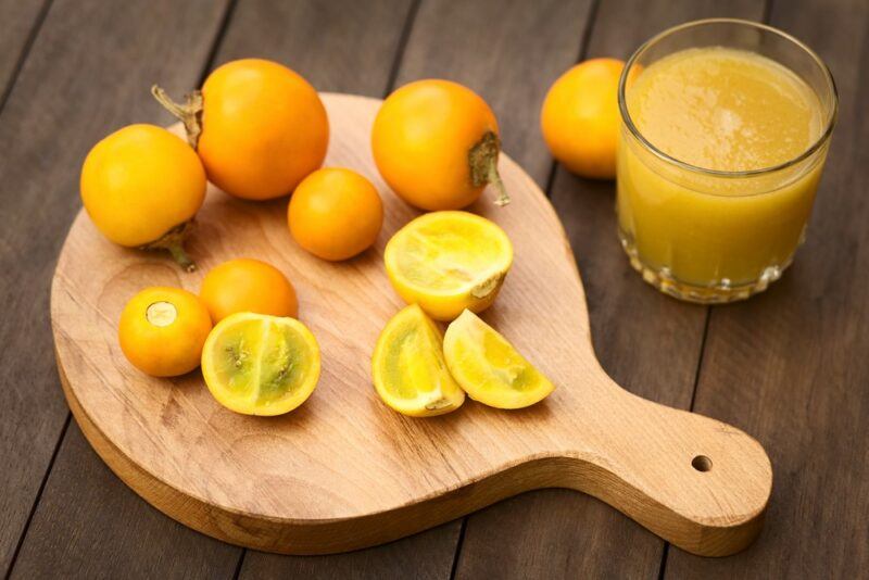 A wooden tray with a collection of small yellow naranjilla fruits, including some that have been cut in half, next to a glass of naranjilla juice