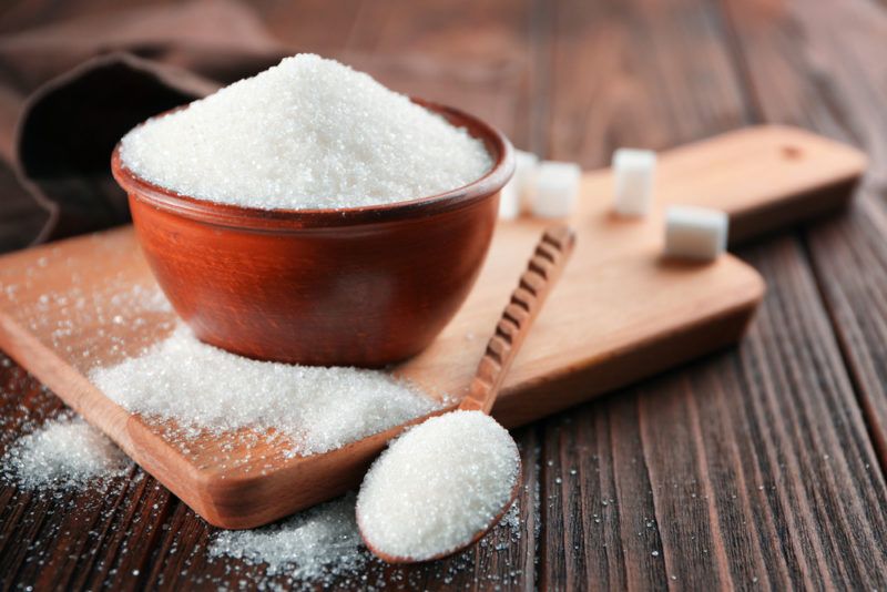 A wooden table with a cutting board that has a bowl of sugar, some sugar cubes, and a spoon