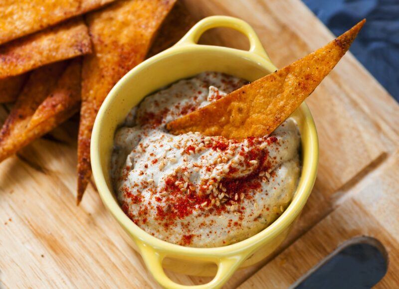 A wooden tray with tortilla chips and a yellow dish of eggplant dip, with one chip being used in the dip