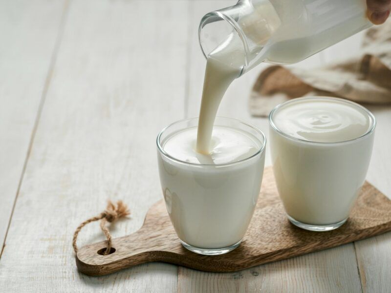 Two glasses of kefir, milk, or drinkable yogurt, one of which is being poured from a bottle.