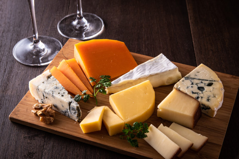 A wooden board of various cheeses next to two glasses of wine