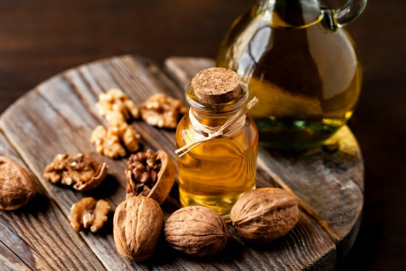 A wooden table with many walnuts, a small jar of walnut oil and a larger jar of the oil