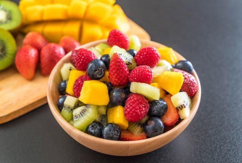 A black table with a wooden bowl of freshly cut fruit, with a black tray with more fruit in the background