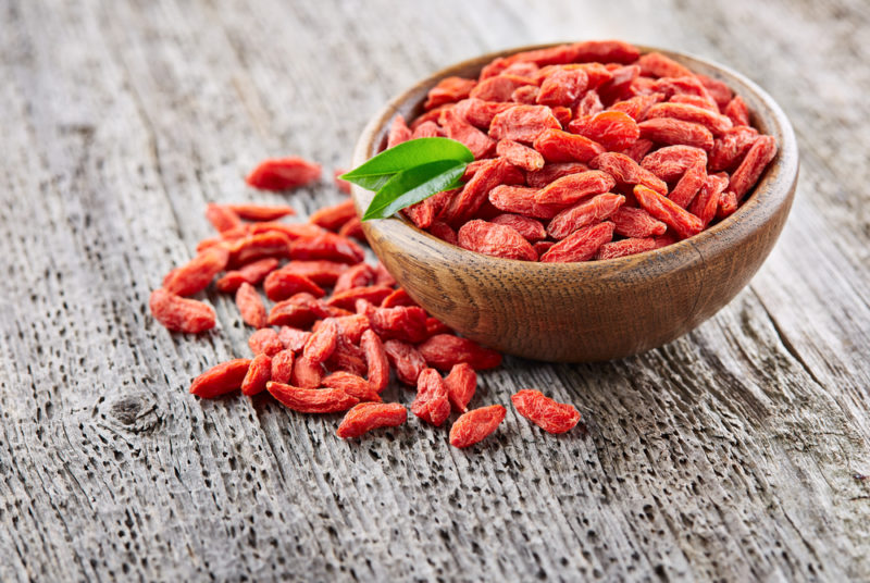 A wooden bowl on a table. There are goji berries in the bowl and more on the table.