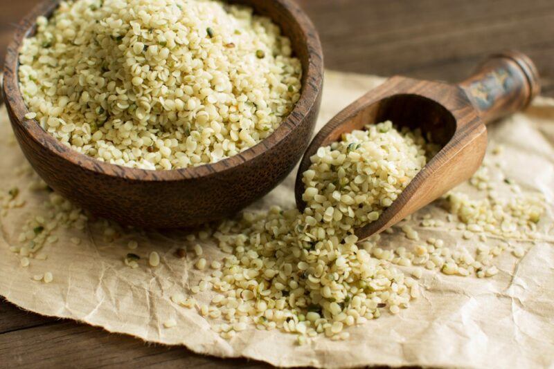 A wooden bowl of hulled hemp seeds, next to a wooden scoop with more seeds, spilling onto paper