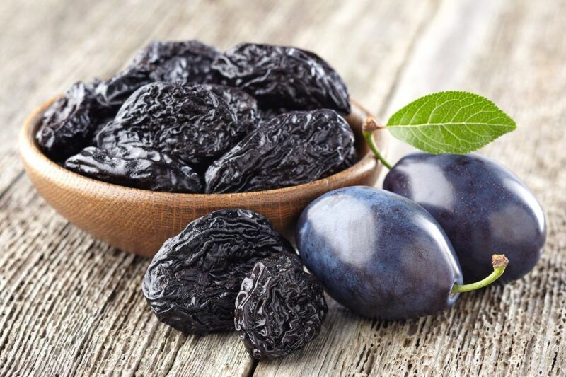 A wooden bowl of prunes, next to two plums