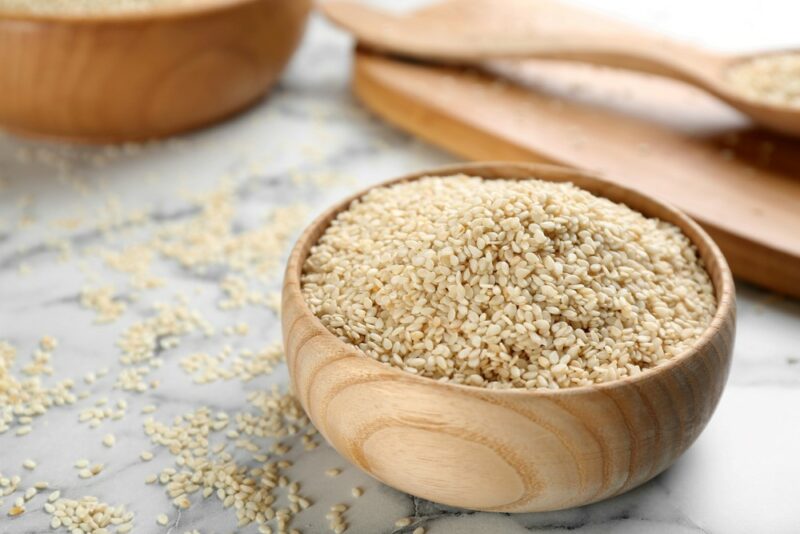 A wooden bowl containing sesame seeds, with more seeds scattered on the table, with wooden items in the background