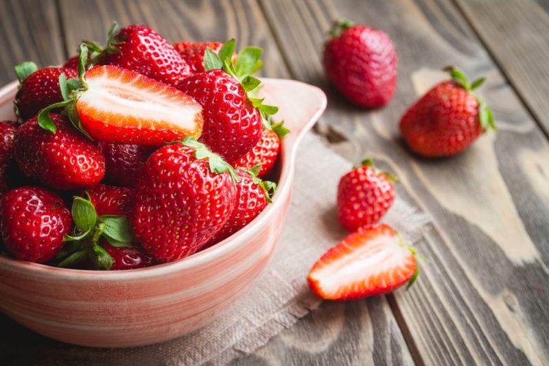 A small bowl filled with strawberries. Most are whole but one is sliced in half. There are a few strawberries on the table too.