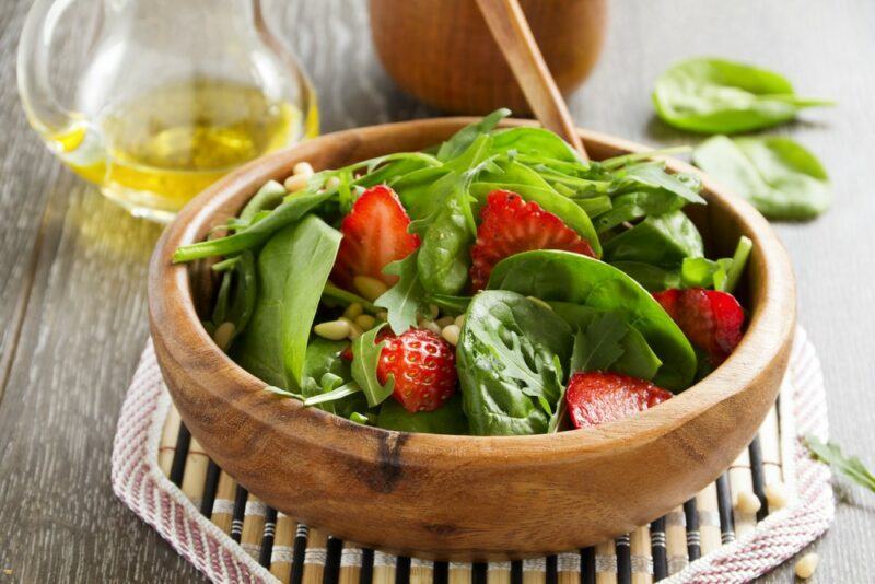 A wooden bowl containing a salad made using spinach and strawberries, with some olive oil in the background