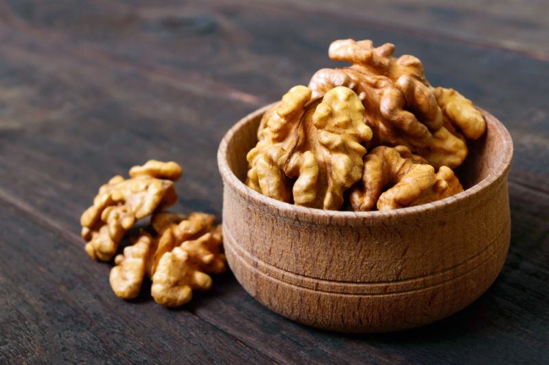 A wooden bowl that contains walnuts with a few extra walnuts on a table