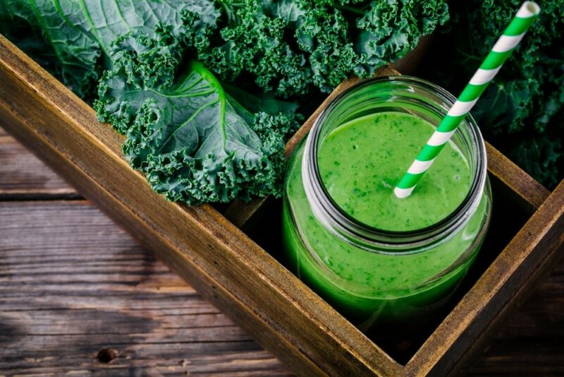 A wooden box containing curly kale, while also containing a green smoothie with a striped straw