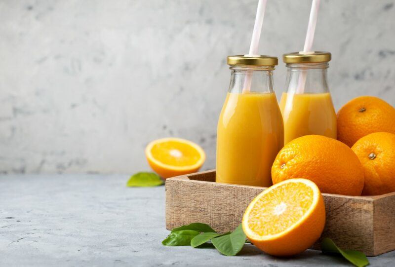 A wooden crate containing orange juice in bottles with straws, plus fresh oranges.