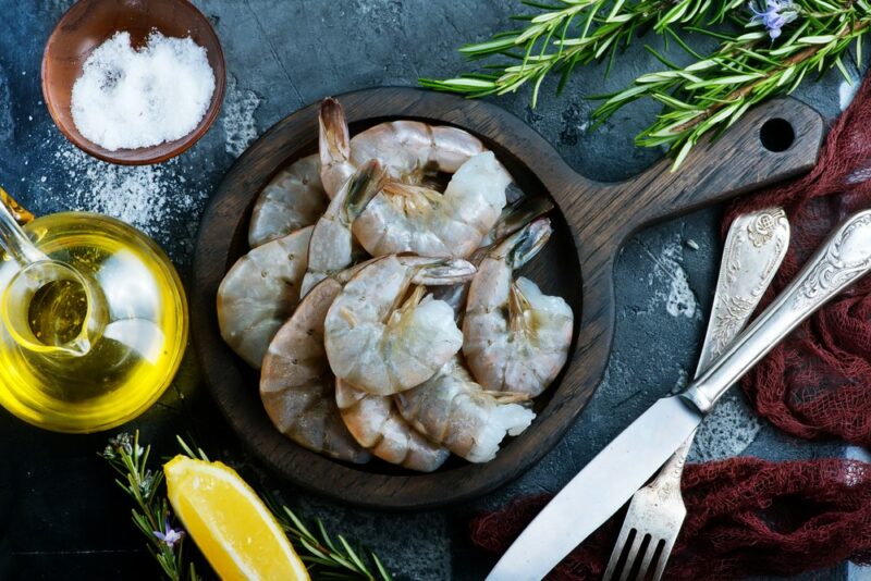 A dark wooden dish filled with raw prawns, with oil, utensils, and salt around the outside