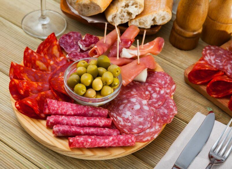 A wooden table with a wooden board that contains a variety of cured meats and a small glass bowl of green olives. There is also a knife and fork, a board with salami, some bread, salt and pepper, and a wine glass on the table