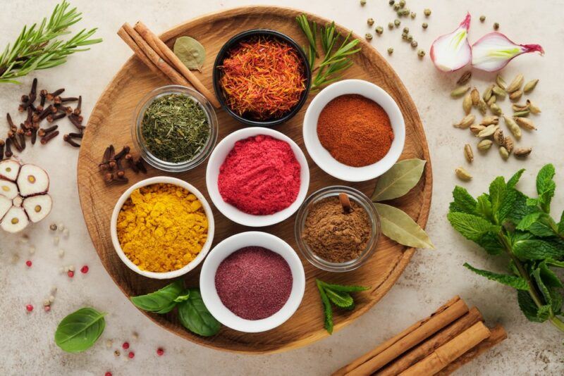 A wooden dish with many small bowls of herbs and spices, with even more herbs and spices scattered on the table.