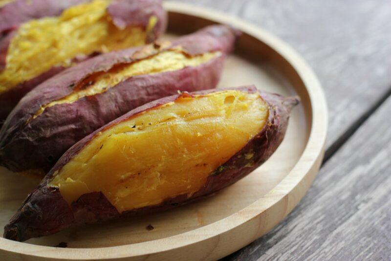 A wooden plate wit three Japanese sweet potatoes. These have been peeled a little so that their yellow flesh can be seen.