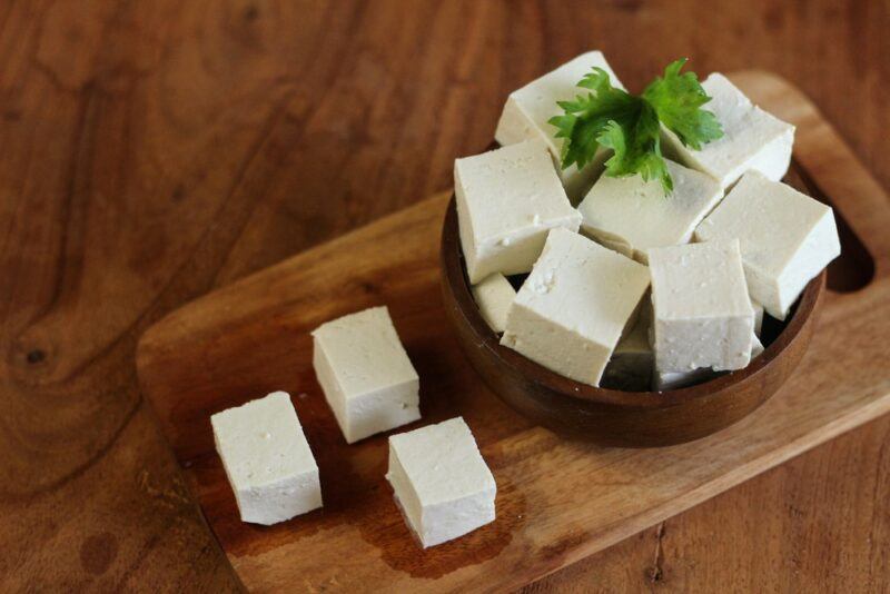 A wooden slab with a bowl of tofu squares and three tofu squares