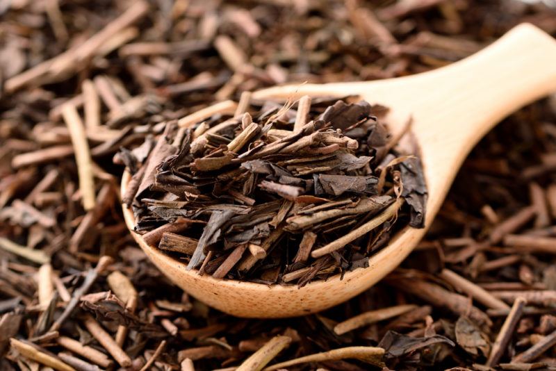 A pile of hojicha sprigs on the ground, with a wooden spoon that contains more of the sprigs