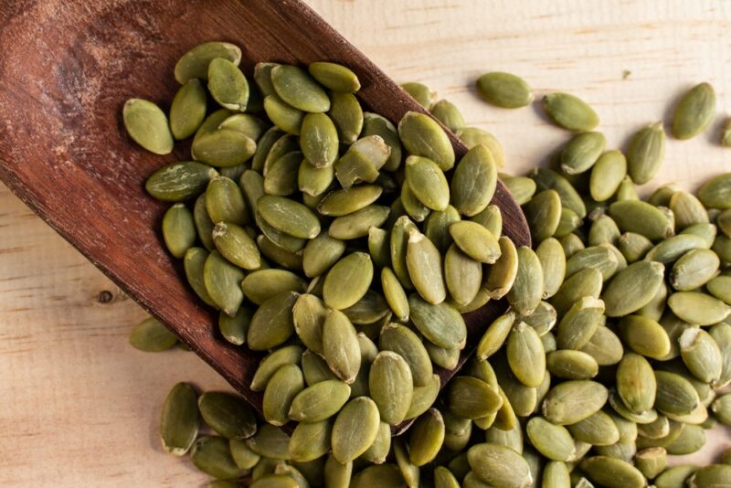 A wooden spoon of pumpkin seeds spilling out onto a table